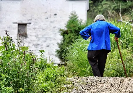Uma mulher idosa segurando uma bengala de costa caminhando em direção a uma casa